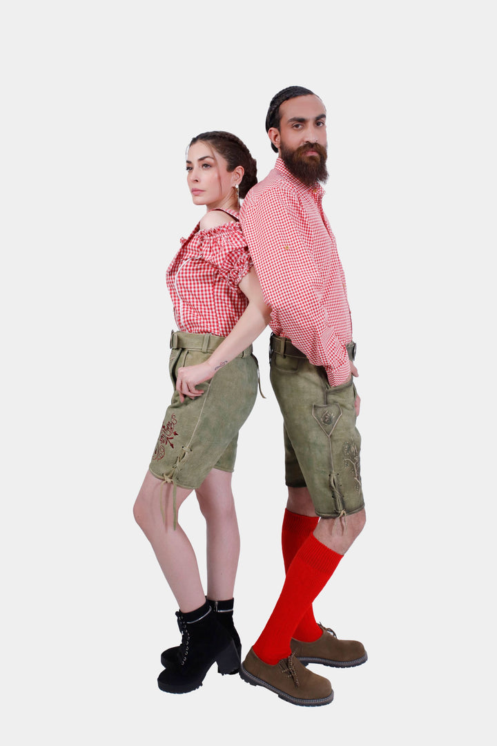 A couple wearing traditional German outfits, standing back-to-back. The woman is dressed in a red and white checkered off-shoulder top with olive embroidered shorts. The man is in a matching red and white checkered shirt, olive embroidered shorts, red knee-high socks, and brown shoes. 