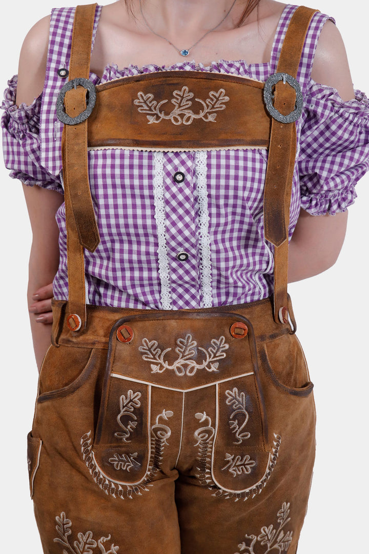 A close-up view of a woman wearing brown leather Angelina Lederhosen with white embroidery and a decorative buckle on the suspenders. She is wearing a purple checkered blouse with off-shoulder sleeves.