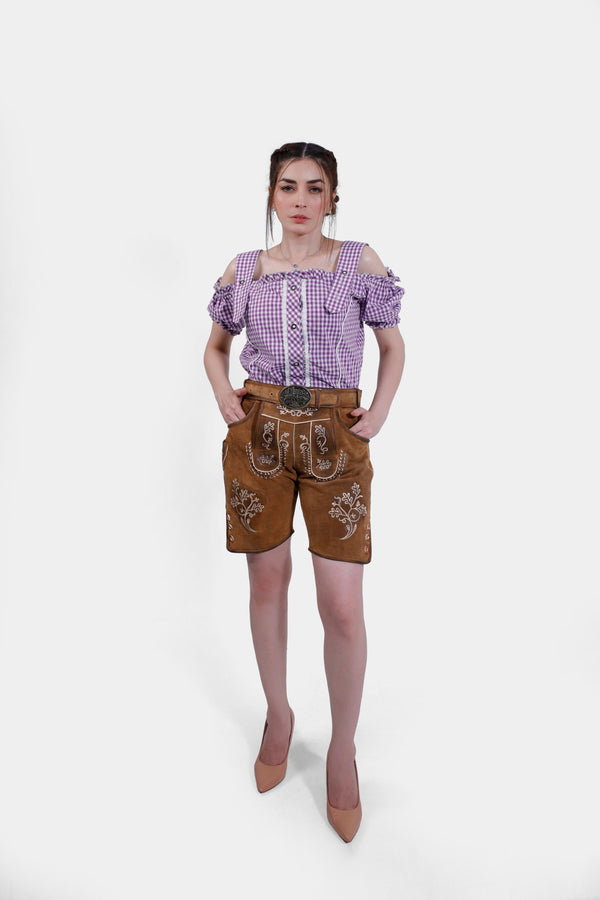 A full-body view of a woman wearing the brown leather Angelina Lederhosen with a decorative belt and white embroidery, paired with a purple checkered blouse. She stands confidently with her hands on her hips and nude high heels. wild lederhose damen braun 