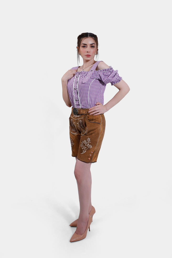 A side view of the woman in the brown leather Angelina Lederhosen, highlighting the side buttons and embroidery. She is wearing a purple checkered blouse and nude high heels.