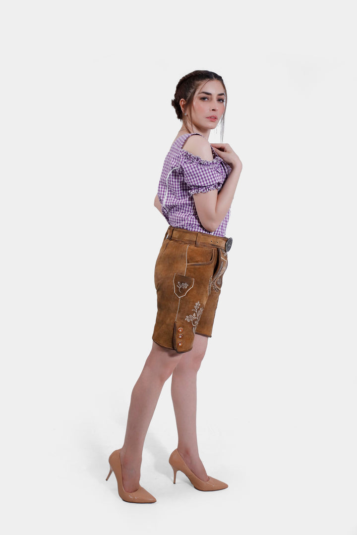 A back view of the woman wearing the brown leather Angelina Lederhosen, showcasing the back design and fit. She is wearing a purple checkered blouse with white detailing and nude high heels.