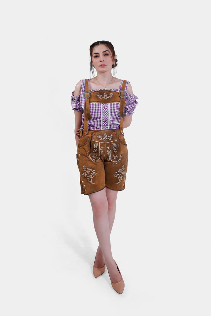 A front view of the woman wearing the brown leather Angelina Lederhosen with suspenders, highlighting the intricate white embroidery. She is paired with a purple checkered blouse with off-shoulder sleeves and stands with her hands on her hips.