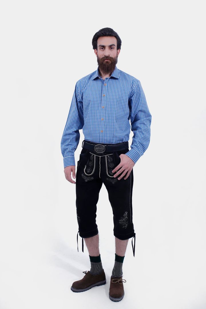 A man with a beard wearing a blue checkered shirt and black Bodensee Bundhosen with traditional socks and brown shoes, standing with his hands by his sides.  Kniebund Lederhose Bodensee mit Träger