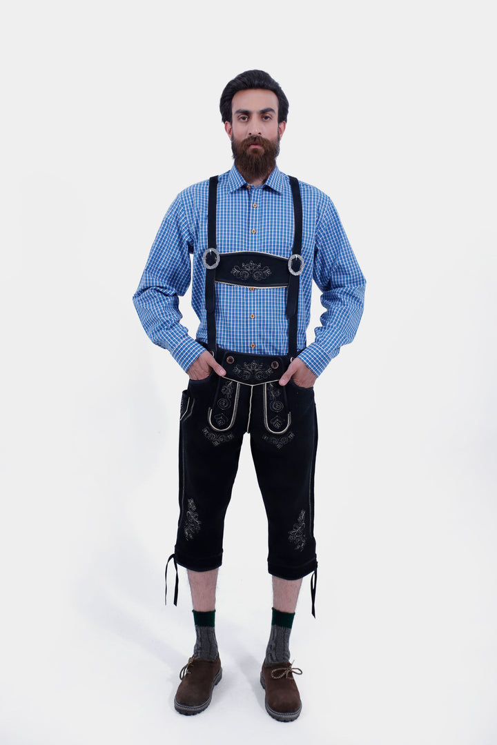  A man with a beard wearing a blue checkered shirt and black Bodensee Bundhosen with suspenders, with traditional socks and brown shoes, posing with his hand in his pocket.