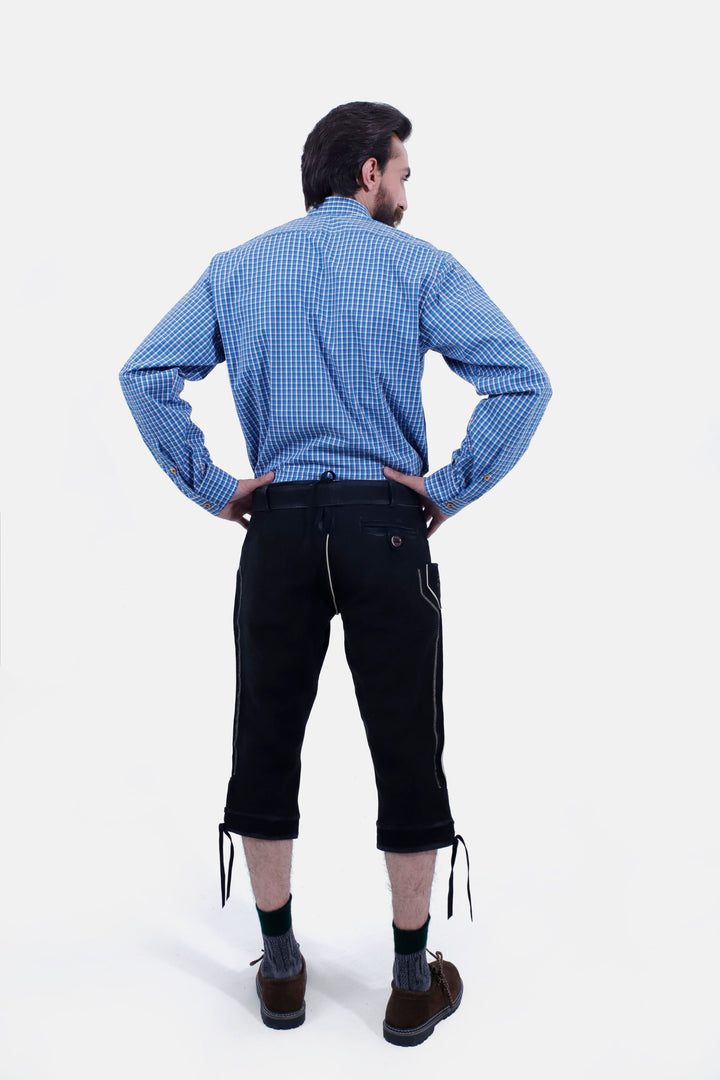 Rear view of a man with a beard wearing a blue checkered shirt and black Bodensee Bundhosen, with traditional socks and brown shoes, with his hands on his hips.