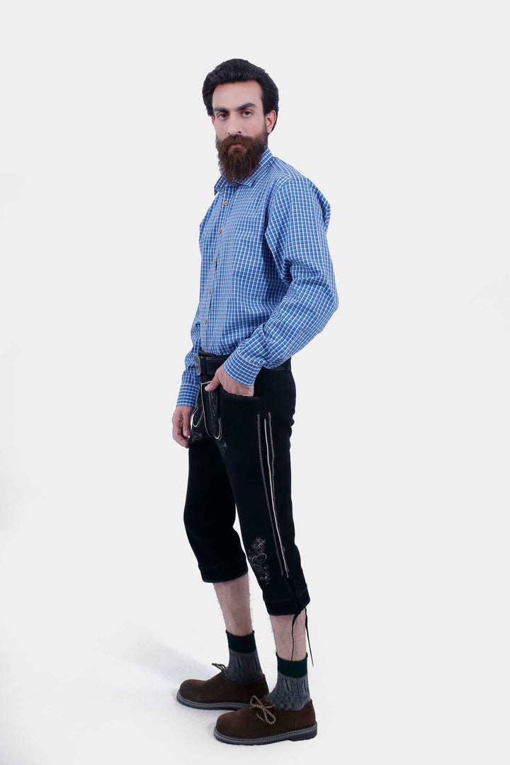 Side view of a man with a beard wearing a blue checkered shirt and black Bodensee Bundhosen, with traditional socks and brown shoes, posing with his hand in his pocket.