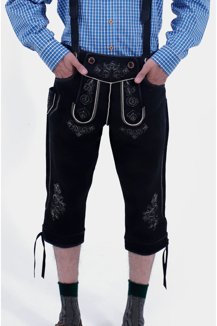 The image shows black leather Bundhosen with white embroidery, worn with a blue checkered shirt. The person has hands in the front pockets and is wearing gray socks with green trim.