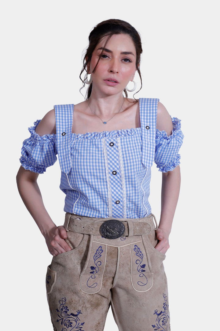 Close-up of a woman wearing a blue and white checkered off-shoulder top with decorative straps and light brown suede lederhosen with intricate blue embroidery