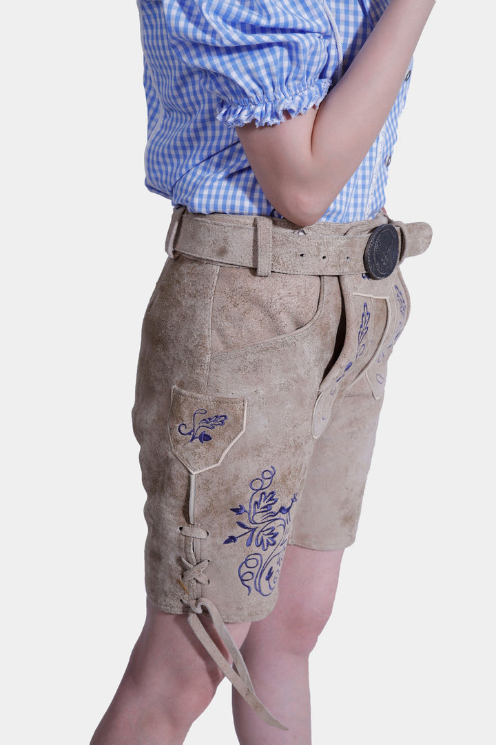 Side view of a woman wearing light brown suede lederhosen with blue embroidery and leather lacing details, paired with a blue and white checkered top.