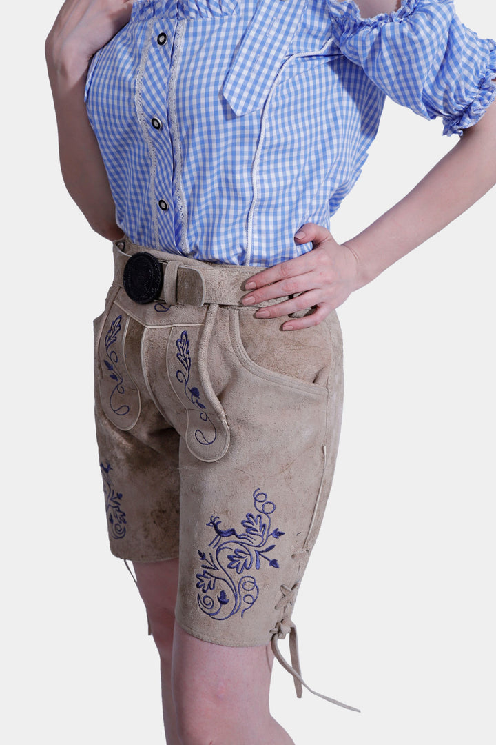 Woman standing with hands above pockets showcasing a blue and white checkered off-shoulder top and light brown suede lederhosen with blue embroidery.