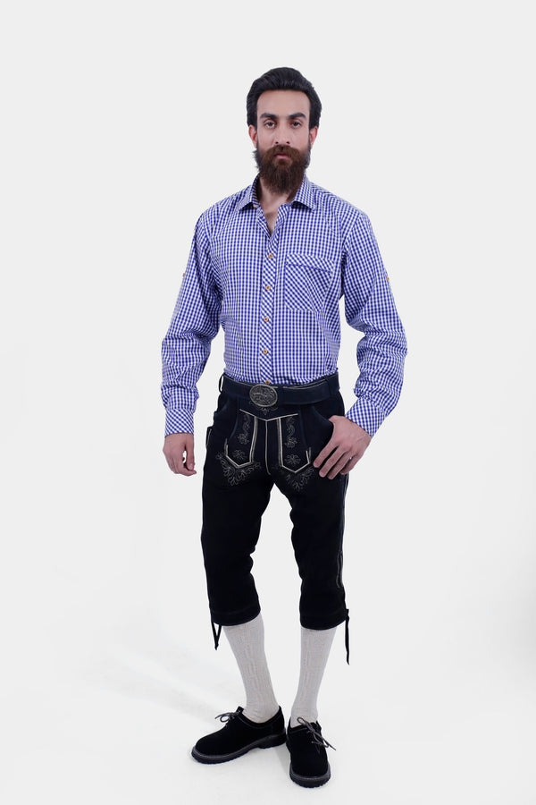 A male wearing a blue checkered shirt and Brandburg Bundhosen Outfit. The Marx Bundhosen features intricate embroidery on the front, a decorative belt buckle, and mid-length. The model's hands are by his sides, and he is wearing black shoes with socks.