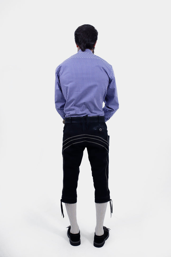 A man with a beard is wearing a blue checkered shirt and Brandburg Bundhosen Outfit with suspenders. The suspenders have decorative buckles and are attached to the pants, which feature intricate front embroidery. The man stands with both hands in his pockets, wearing black shoes and socks.