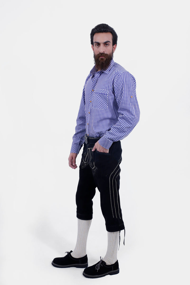 A male standing in a semi-profile pose, wearing a blue checkered shirt and Brandburg Bundhosen Outfit. One hand is in his pocket, showcasing the side embroidery, small pocket details, and overall fit of the pants. With traditional Bavarian embroidery and a decorative belt. He is wearing black shoes and socks