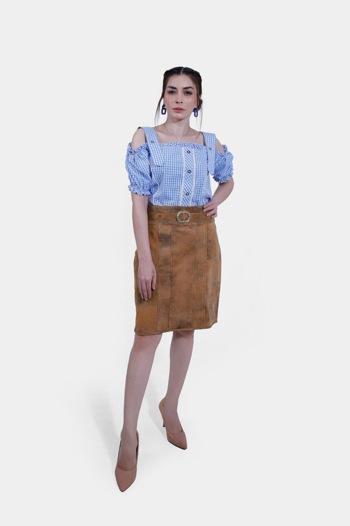 Close-up view of a Clara Lederhosen skirt, showcasing the detailed texture of the brown leather material and the decorative belt buckle, with the woman crossing her arms over her blue and white checkered blouse. lederrock midi