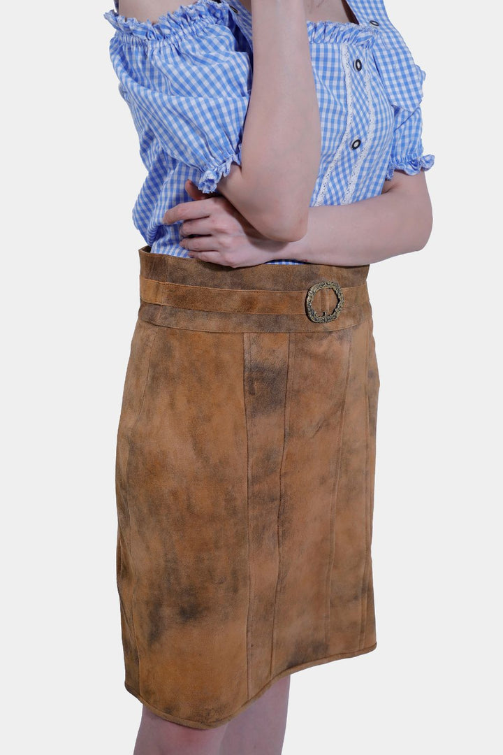 A woman is posing in a Clara Lederhosen skirt and a blue and white checkered off-shoulder blouse, with her hand resting on her chin, standing against a plain white background.