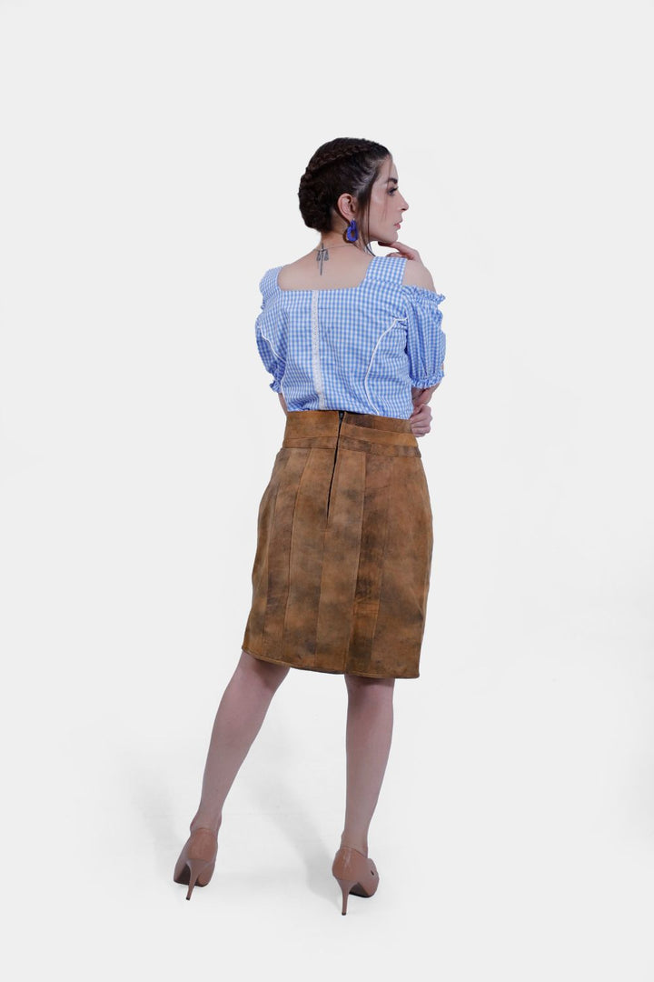 A woman is standing with her hands on her hips in a Clara Lederhosen skirt and a blue and white checkered off-shoulder blouse, highlighting the detailed texture of the brown leather skirt and its decorative belt buckle, against a plain white background.