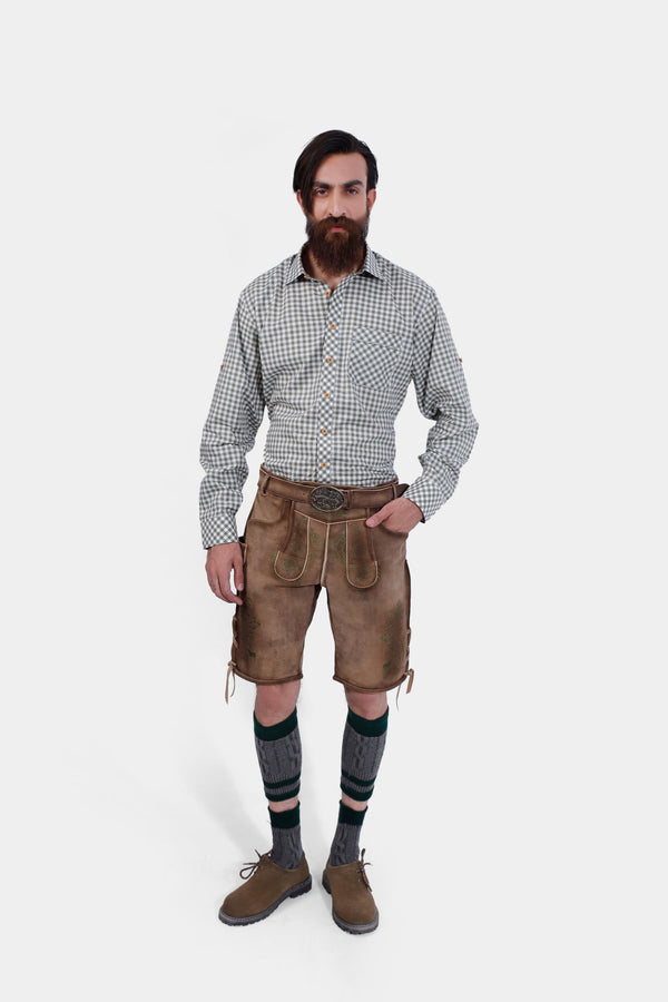 Full body view of a man wearing a green and white checkered shirt and brown leather lederhosen with knee-high socks and brown shoes, standing with a serious expression. lederhose braun herren​, lederhose herren braun