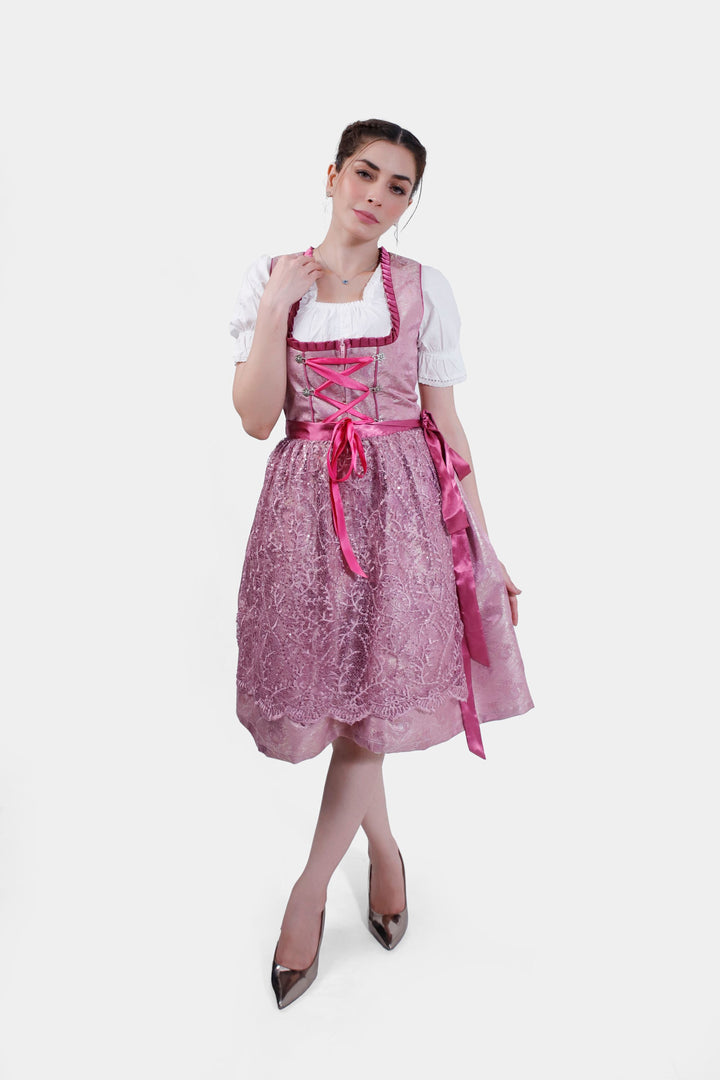 Full-length view of a woman wearing the Dirndl Christiane, showcasing its shimmering pink lace skirt and satin apron tied at the waist.