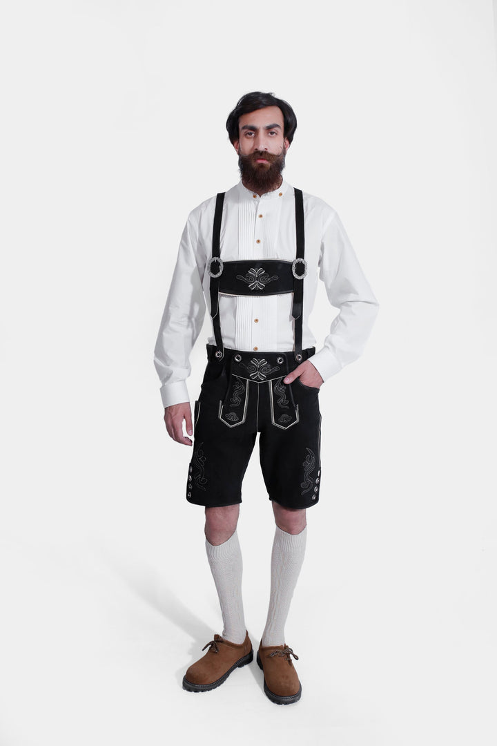 A man with a beard wearing the Elgort Lederhosen Outfit, which includes a white shirt, black leather Lederhosen with white embroidery and suspenders, white knee-high socks, and brown shoes. lederhose herren schwarz tracht​ 