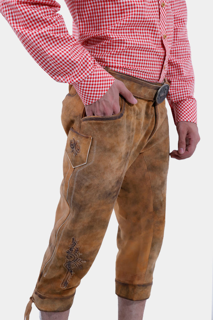 Side view of tan brown Eltz Bundhosen featuring embroidery on legs and lases on cuffs, worn by a man against a white background.