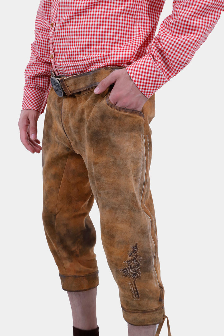 Side view of tan brown Eltz Bundhosen featuring embroidery on legs and lases on cuffs, worn by a man against a white background.