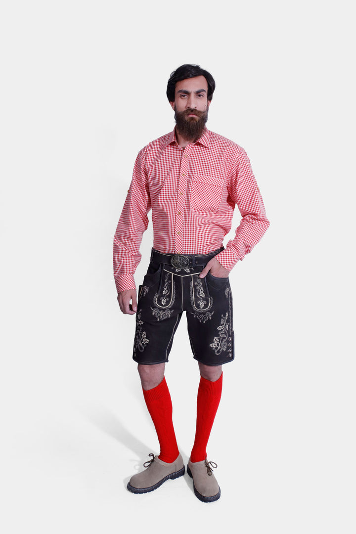 Man wearing an Everglades Lederhosen Outfit, featuring a red and white checkered shirt, black leather lederhosen with intricate white embroidery, traditional red knee-high socks, and leather shoes. trachten lederhose schwarz herren , schwarze lederhose tracht