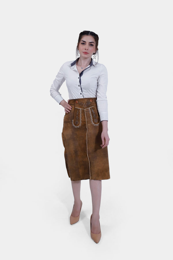 Woman wearing the Griessee Lederhosen Skirt in brown leather paired with a white button-up shirt, standing with one hand on her hip and the other by her side. lederrock knielang, lederrock braun knielang