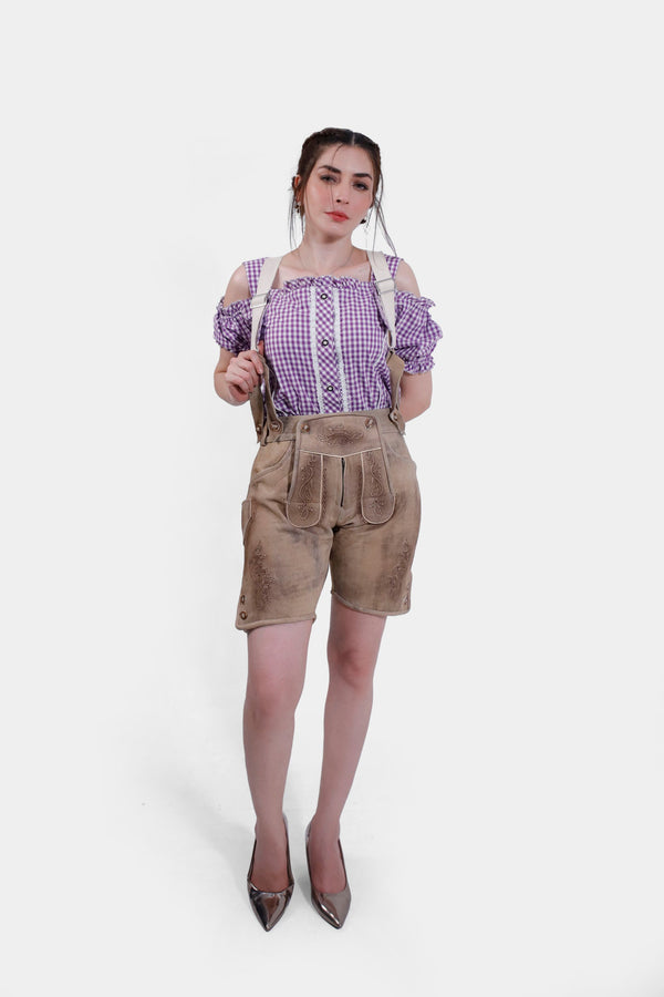 A full-body view of a woman wearing the tan leather Hamburg Lederhosen with suspenders, paired with a purple checkered blouse. She stands confidently with her hand on her hip and metallic high heels. braune lederhose damen