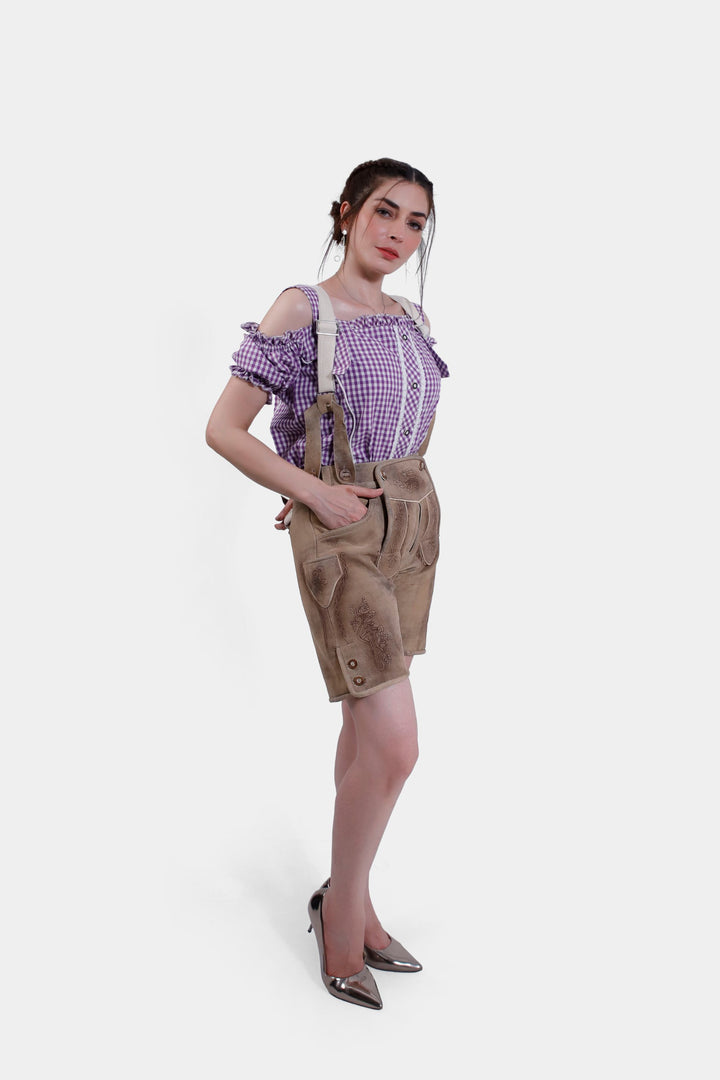 A side view of the woman in the tan leather Hamburg Lederhosen, highlighting the embroidery and side buttons. She is wearing a purple checkered blouse and metallic high heels.