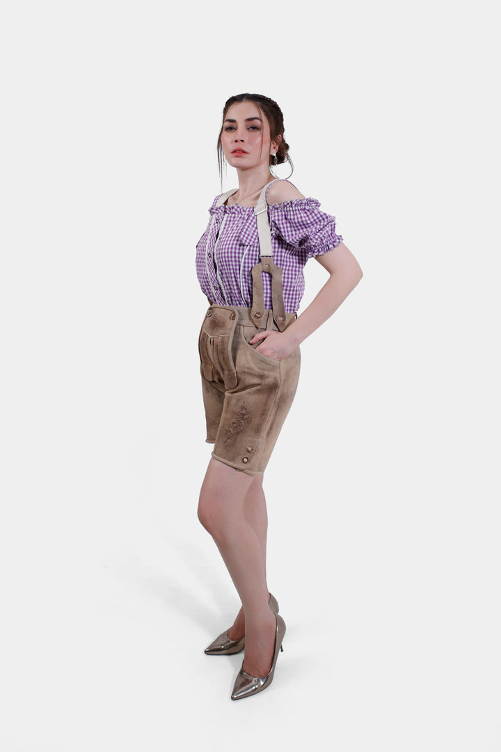 A full-body shot from a different angle, showing the woman in tan leather Hamburg Lederhosen with suspenders and a purple checkered blouse. She poses with one hand in her pocket and metallic high heels.