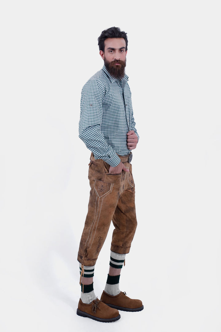 Man dressed in a green checkered shirt and Hesse Bundhosen, posing with a slight side view. The brown leather pants feature intricate floral embroidery, side pockets, and are complemented by traditional Bavarian shoes and green-striped socks.