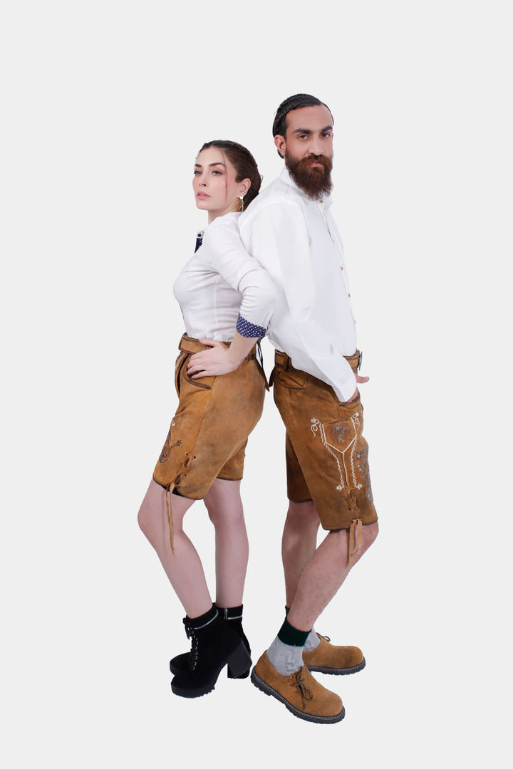 A couple in traditional German clothing standing back-to-back. The woman is in a white blouse with blue accents and tan leather shorts with floral embroidery. The man is wearing a white button-down shirt and tan leather shorts with floral embroidery, green socks, and brown leather shoes. Both are looking serious, standing with arms by their sides.