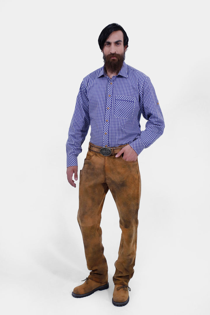 A full-body front view of a bearded man with black hair wearing a blue and white checkered shirt tucked into Humboldt Long Lederhosen, posing with one hand in his pocket and looking at the camera. lederhose tracht herren lang