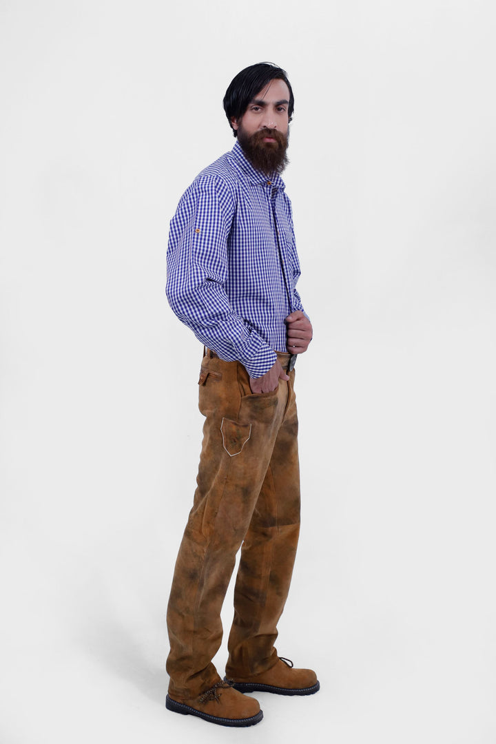 A full-body side view of a bearded man with black hair wearing a blue and white checkered shirt tucked into Humboldt Long Lederhosen, standing with his hands in his pockets.