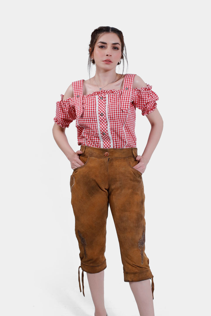 A woman is wearing a red and white checkered, off-shoulder blouse with ruffled sleeves and the "Kirsten Dunst Lederhosen" brown leather pants that end just below the knee. She is standing with her hands in her pockets and looking directly at the camera.