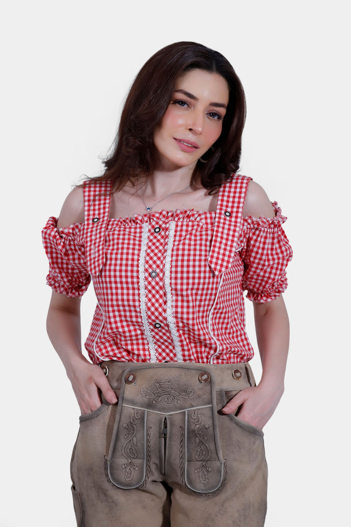 A woman wearing a red checkered off-shoulder blouse and traditional brown leather lederhosen, standing with her hands in her pockets, looking at the camera.