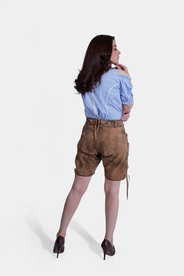 Full body shot of woman in Lorelei Lederhosen, standing with crossed legs and hands in pockets, featuring blue checkered blouse.