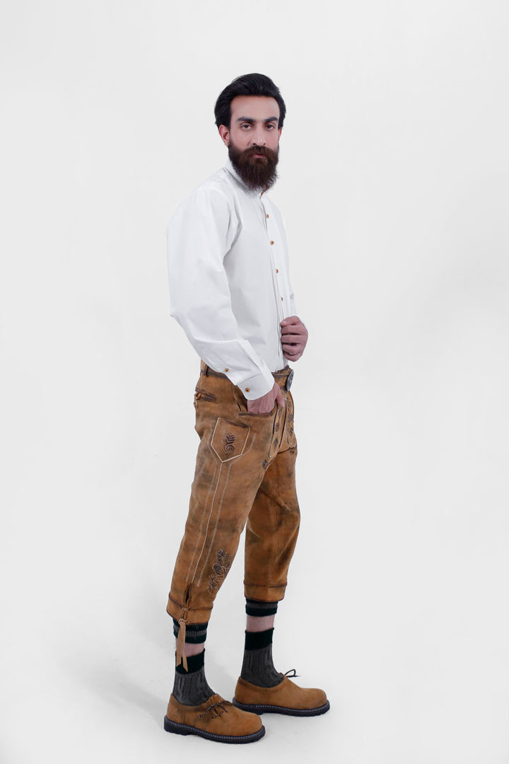 A side view of a man wearing Lothar Bundhosen Outfit. Wearing white traditional shirt, brown lederhosen with intricate designs, dark socks, and traditional brown shoes.