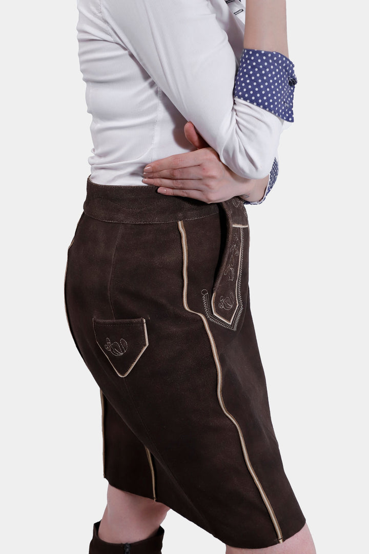 Side view of a woman wearing the Louise Lederhosen Skirt in dark brown leather, highlighting the skirt's texture, front flap with embroidery, and side pocket detail.