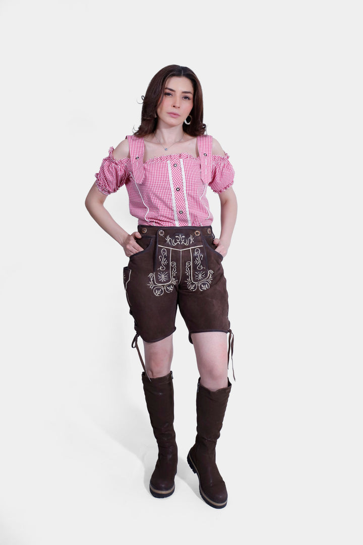 Woman standing confidently in traditional brown Margarethe Lederhosen and pink checkered blouse, detailed with intricate white floral embroidery. braune lederhose, Damen Trachten Lederhose Kurz