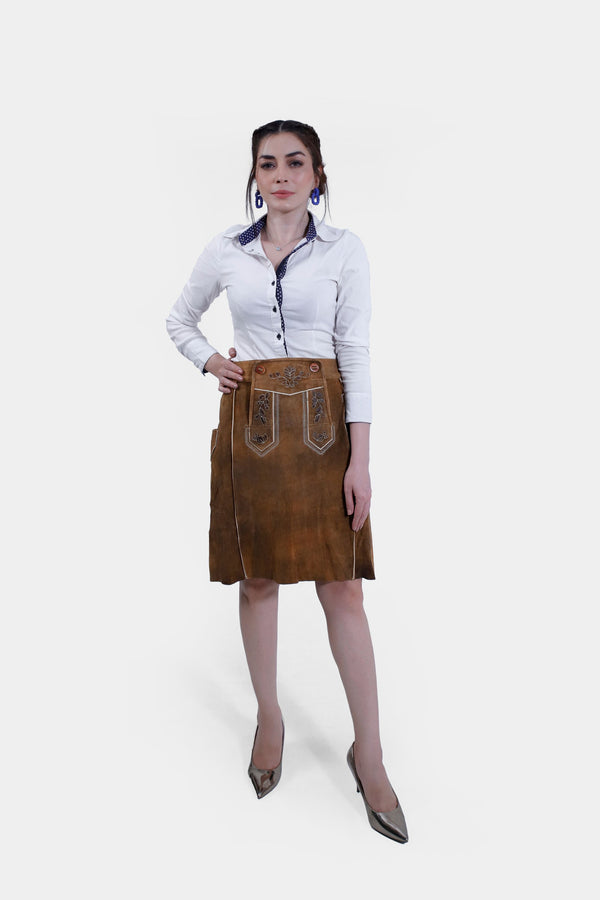 A woman wearing the Marianna Lederhosen Skirt, front view. The skirt is made of brown leather with intricate embroidery, paired with a white button-down shirt. lederrock braun,  lederröcke knielang