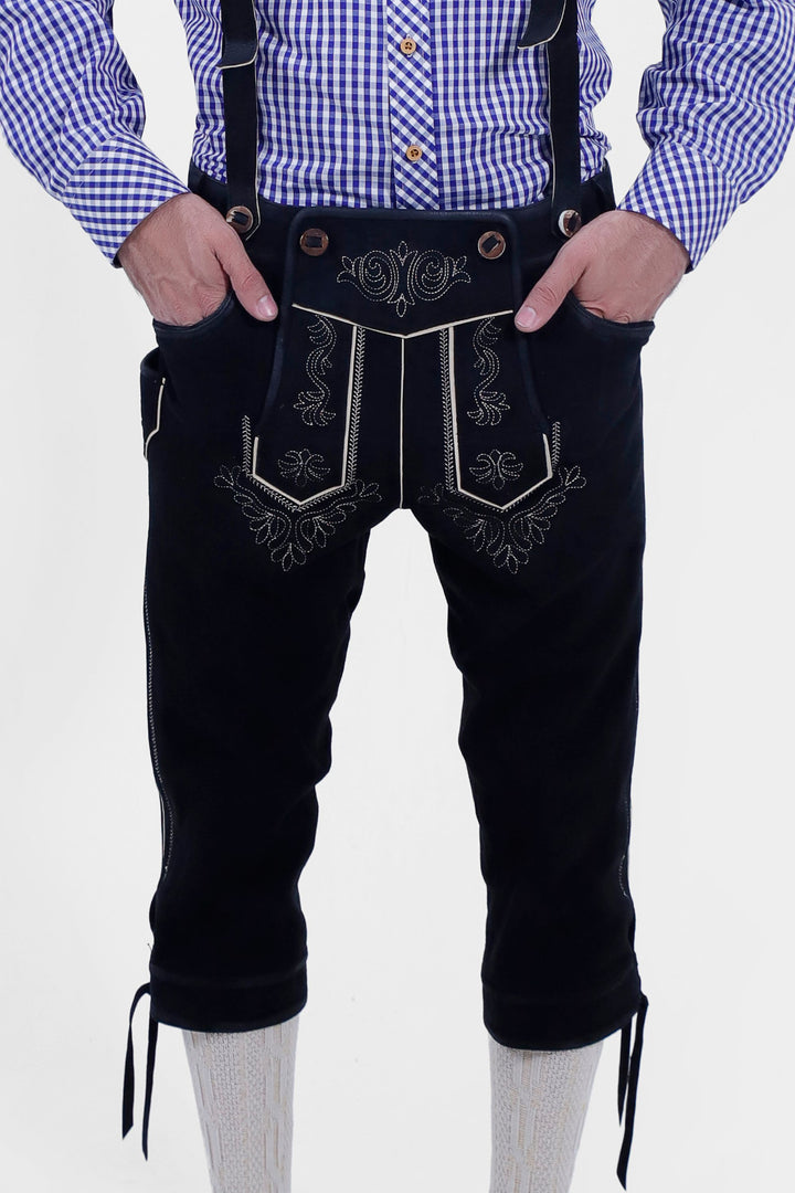 The image shows a pair of traditional black leather Marx Bundhosen with white embroidery, worn with a blue and white checkered shirt. The Bundhosen have intricate designs on the front and sides, and the person has their hands in the front pockets.