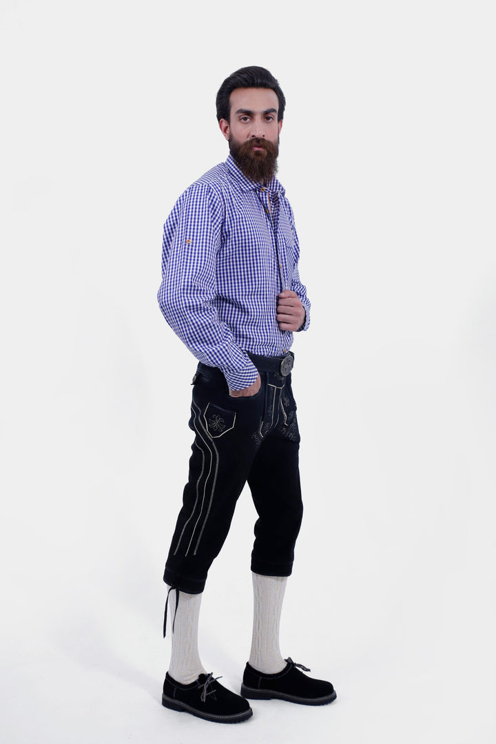 A male shown in a side profile wearing a blue checkered shirt and Marx Bundhosen. One of his hands is in the pocket of the pants, highlighting the side embroidery and detailing, including a small side pocket. The model is wearing brown shoes with socks, and the mid-length pants are secured with a decorative belt.