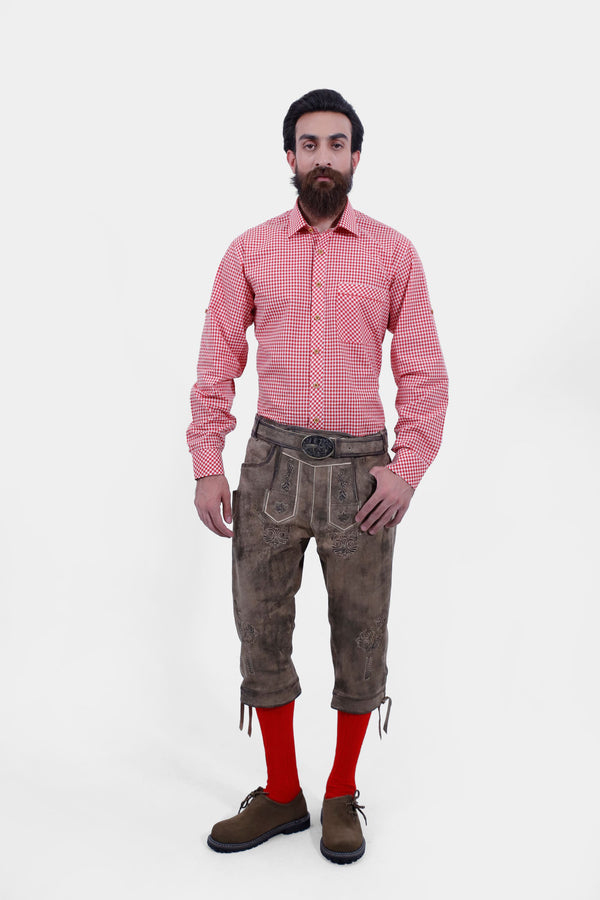 A male wearing a red checkered shirt and Milwaukee Old World Bundhosen. The Milwaukee Old World Bundhosen features intricate embroidery on the front, a decorative belt buckle, and mid-length. The model's hands are by his sides, and he is wearing brown shoes with red socks. herren lederhose kniebund, trachten lederhose kniebund herren