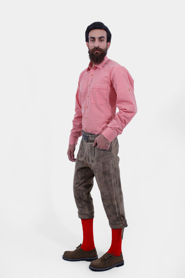 A side view of a man wearing Close-up side view of Milwaukee Old World  Bundhosen. He is in a red checkered shirt, red socks, and brown shoes, with his hand in his pocket.