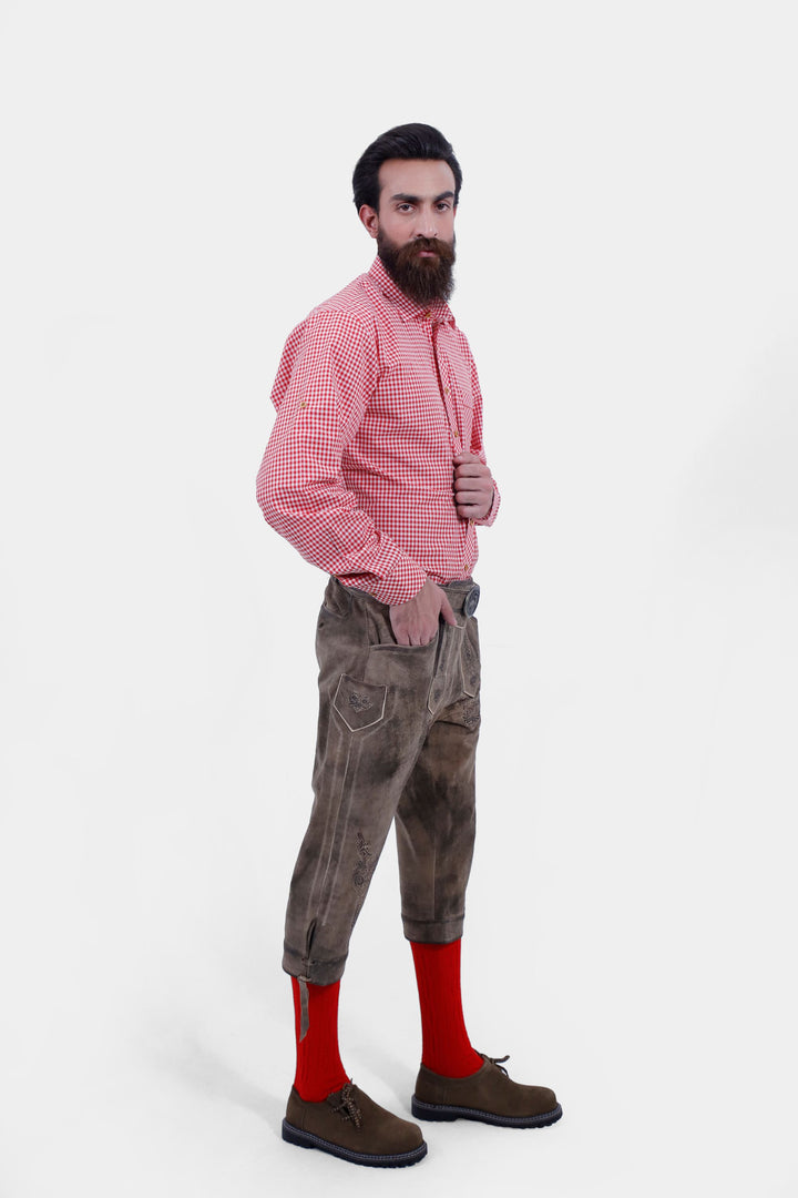 A man posing in  Milwaukee Old World  Bundhosen. He is dressed in a red checkered shirt, knee length lederhosen, red socks, and brown shoes, with his hand near his chest.