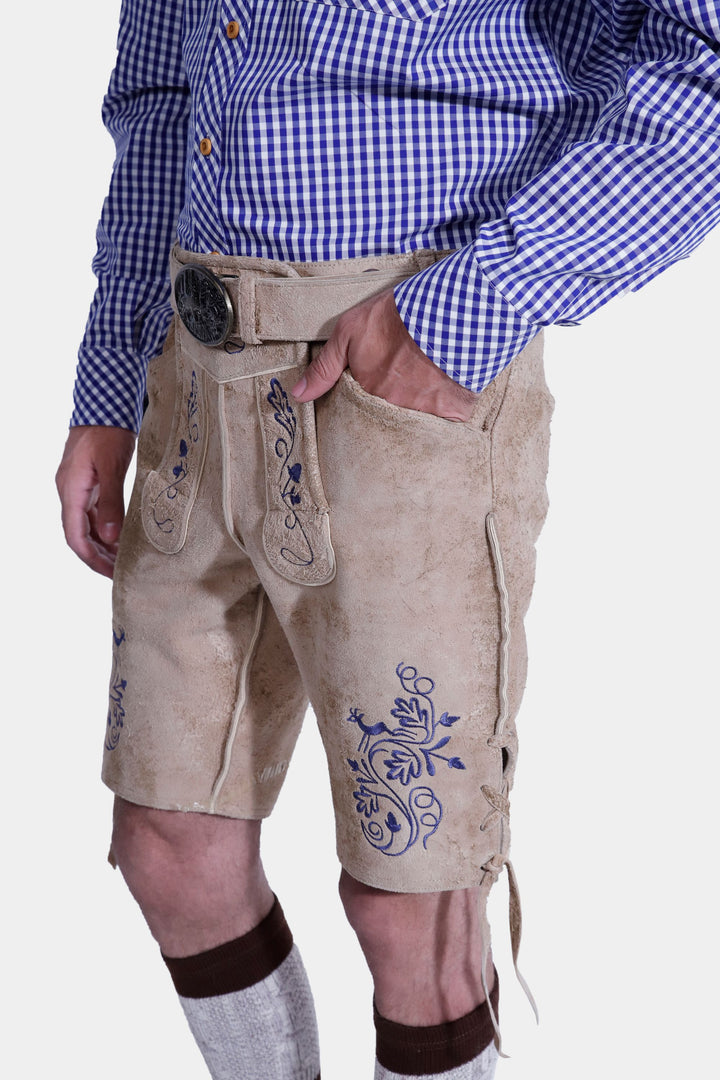 Side view of the Mookie Lederhosen Outfit, focusing on the detailed blue embroidery on the tan leather lederhosen, paired with a blue and white checkered shirt and hand in the pocket.