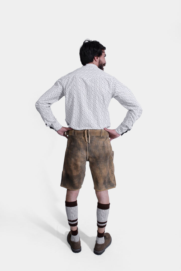 Rear view of a man wearing Mozart Lederhosen, highlighting the back pockets and detailed stitching of the traditional Bavarian leather shorts. He is also wearing a white patterned shirt and beige shoes.