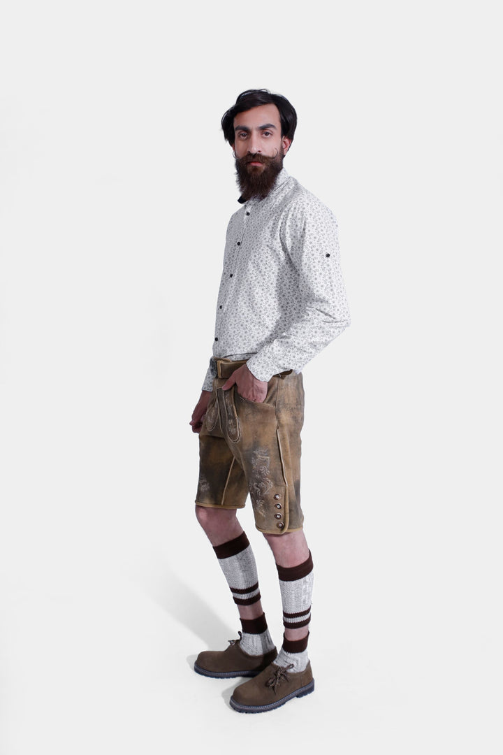 Side profile of a man in Mozart Lederhosen, emphasizing the leather shorts' design, embroidery, and fit, paired with a white patterned shirt and beige shoes.