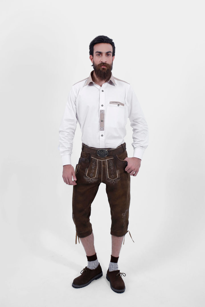 A male wearing a white shirt and Oslo Bundhosen. The Oslo Bundhosen features intricate embroidery on the front, a decorative belt buckle, and mid-length. The model's hands are by his sides, and he is wearing brown shoes with grey socks. herren kniebundlederhose, kniebundlederhose herren